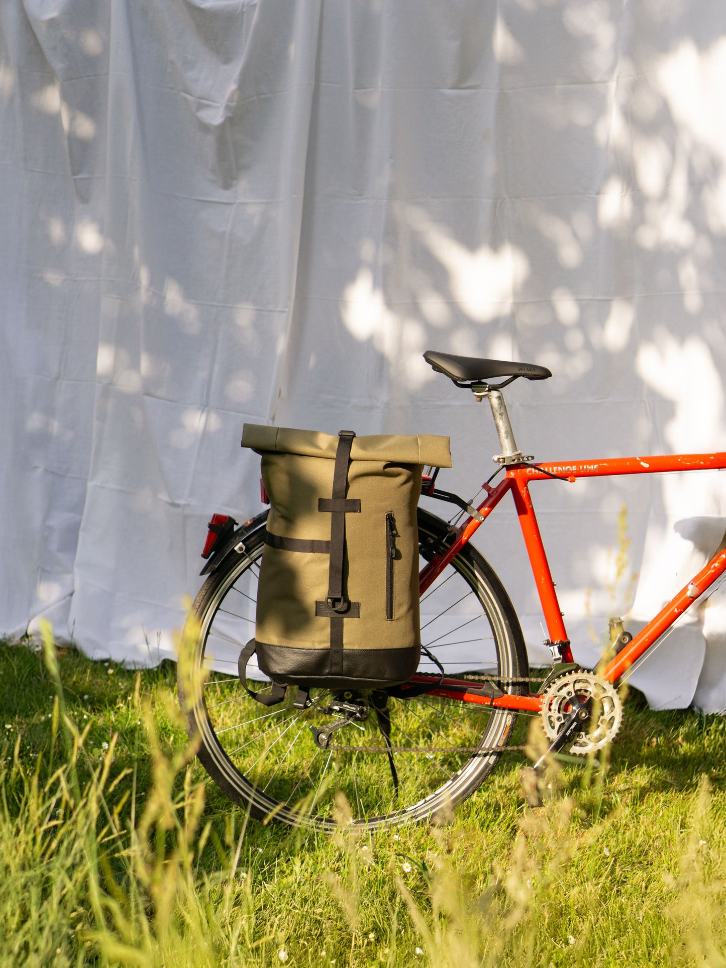 Ein rotes Fahrrad steht auf einer grünen Wiese, am Gepäckträger ist der Fahrradrucksack Fixie Olive von Spleen Berlin befestigt. Der urbane Rucksack vereint Stil und Funktion, ideal für Pendler:innen nachhaltig produziert und mit praktischer Fahrradhalterung ausgestattet. ©Spleen Berlin
