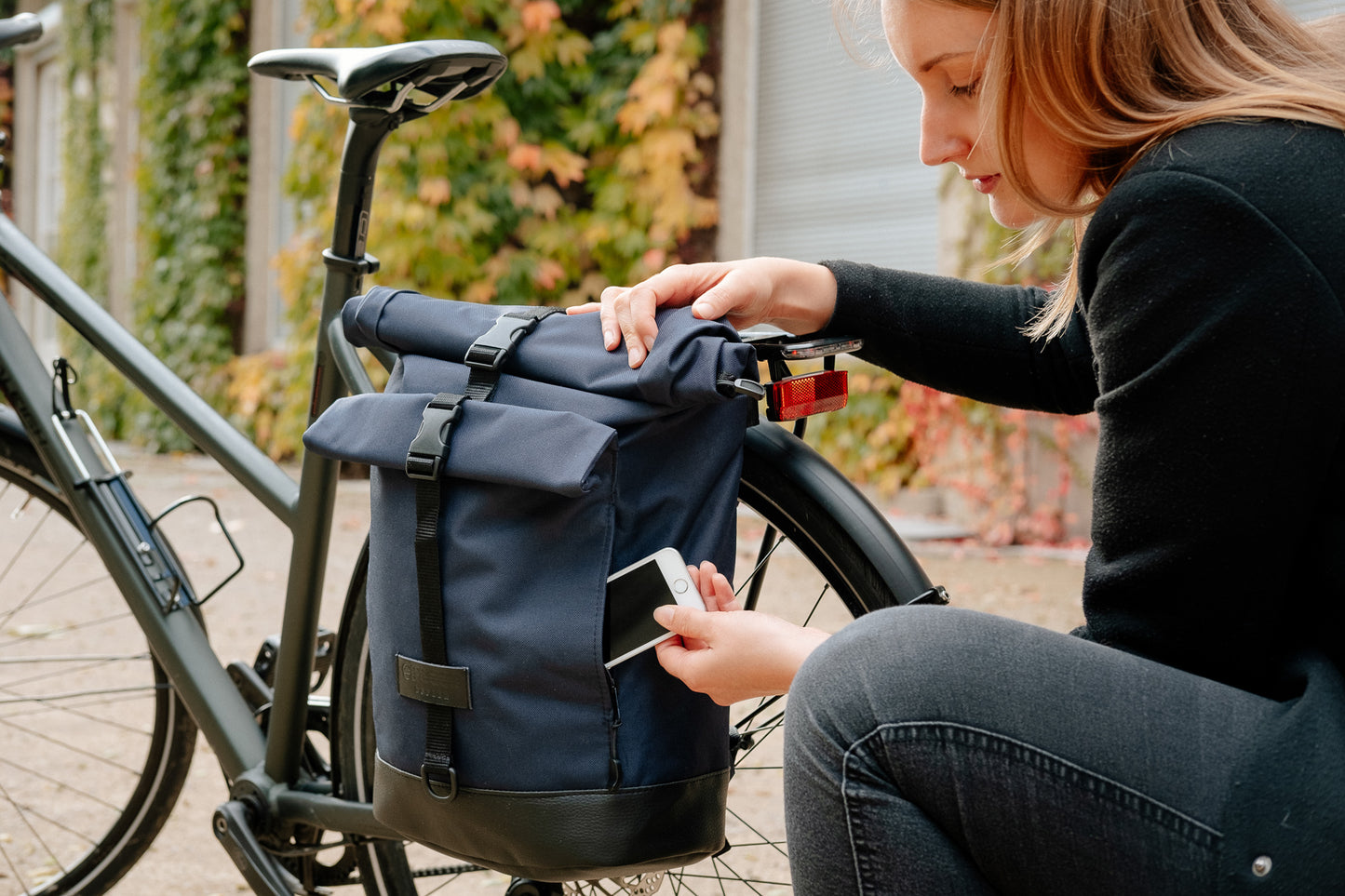 BOB Fahrradtasche in Marineblau von Spleen Berlin am Gepäckträger befestigt, Frau•kniet daneben und nimmt ihr Handy aus der Fronttasche; vegan & hybrid nutzbar. ©Spleen Berlin